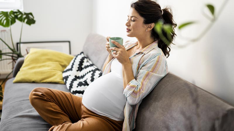 Mulher grávida sentada em sofá de pernas cruzadas, segura numa caneca com as duas mãos junto ao rosto e parece inspirar o aroma de olhos fechados.